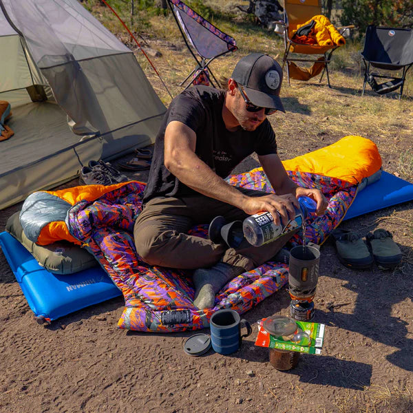 Man camping, sitting on a colorful sleeping pad with a tent and chairs in the background.