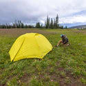 Yellow tent set up in a grassy field with a person sitting nearby under a cloudy sky.