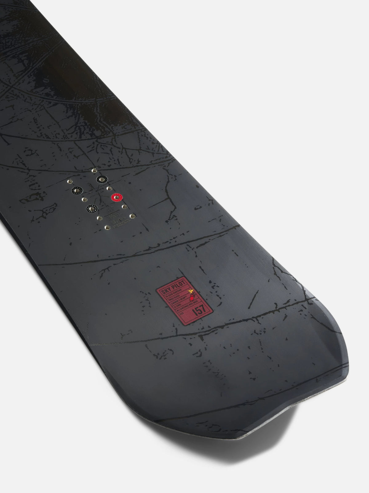 A close-up of a snowboard base showing the underside with a dark gray and black map-like graphic pattern, a red logo patch, and binding device eyelets, photographed on a light gray background.
