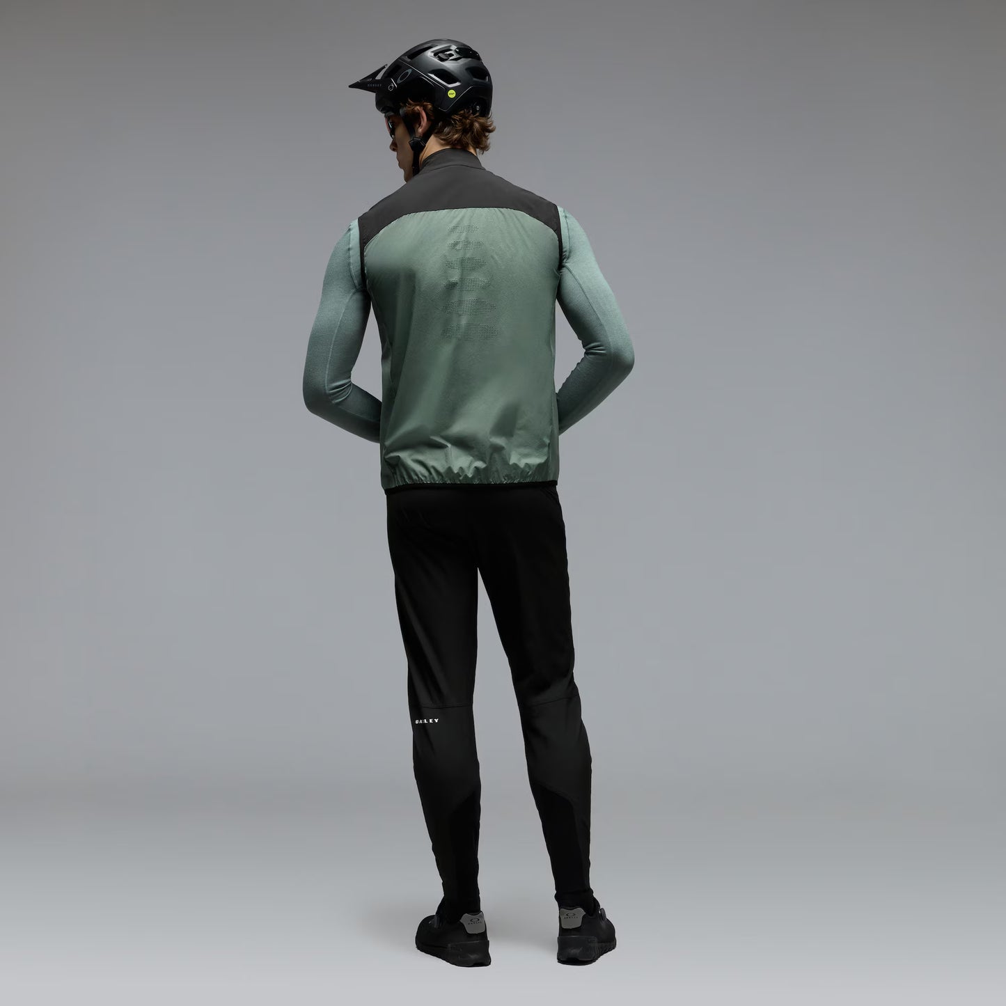 A man wearing a green and black cycling vest over a long-sleeve green shirt, black cycling pants, and a black helmet, shown from the back against a gray studio background.