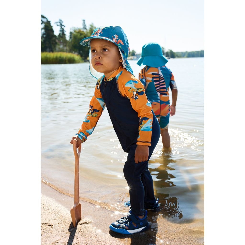 Two children in wetsuits and hats playing in water near a forest.