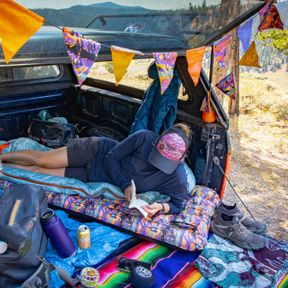 Person lying on a colorful sleeping pad in the back of a vehicle with camping gear and flags.