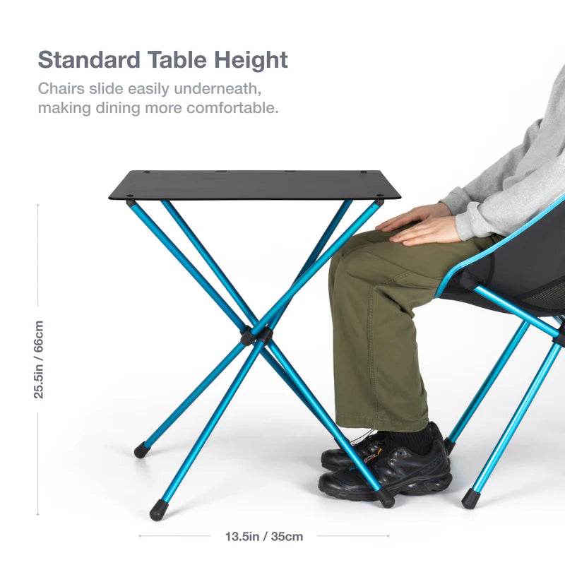 Portable folding table with blue legs and black top, shown with a person sitting on a chair underneath.