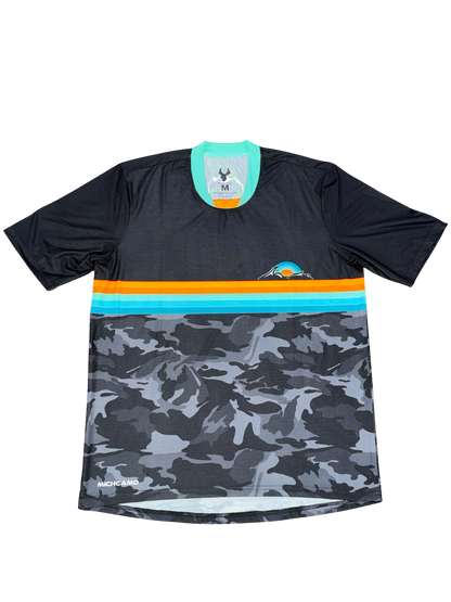 A short-sleeve athletic jersey with a black upper section, a horizontal stripe band in orange and turquoise, and a gray/black camouflage pattern on the lower half. The brand name 'Machete Apparel' is visible near the bottom left hem.