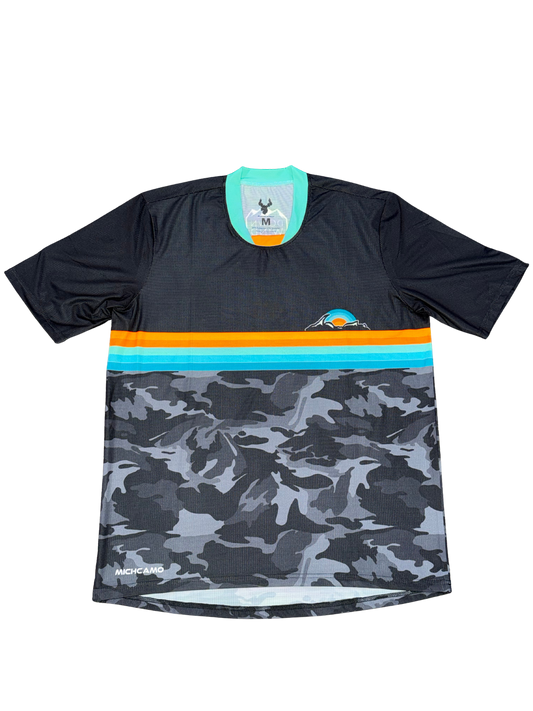 A short-sleeve athletic jersey with a black upper section, a horizontal stripe band in orange and turquoise, and a gray/black camouflage pattern on the lower half. The brand name 'Machete Apparel' is visible near the bottom left hem.