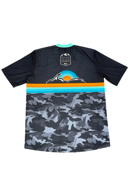 A short-sleeve athletic jersey shown from the back, featuring a dark navy upper section with a mountain and sunset graphic, a horizontal stripe band in teal, orange, and light blue, and a gray and black camouflage pattern on the lower half. The brand 'DOWN WIND SPORTS' is printed on the back neck patch.