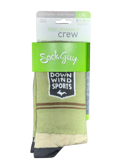 A pair of SockGuy crew socks in a light olive green colorway, featuring a black and white 'Down Wind Sports' shield patch on the side, with brown stripes and a beige cuff. The socks are shown in their retail packaging against a white background.