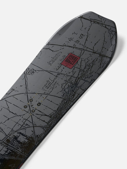 A close-up of a snowboard deck featuring a dark gray and black topographic map design with a small red square logo near the center, photographed on a light gray background.