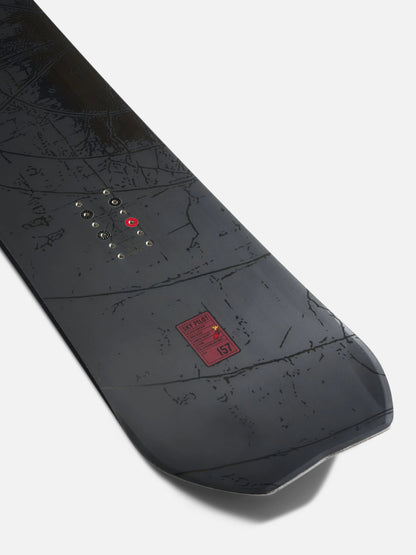 A close-up of a snowboard base showing the underside with a dark gray and black map-like graphic pattern, a red logo patch, and binding device eyelets, photographed on a light gray background.