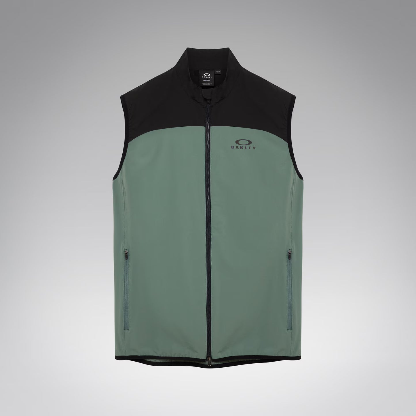 A sleeveless zip-front vest with a black upper yoke, collar, and hem, and a muted sage green lower body. The vest has a small Oakley logo on the left chest and zippered side pockets, shown on a light gray gradient background.