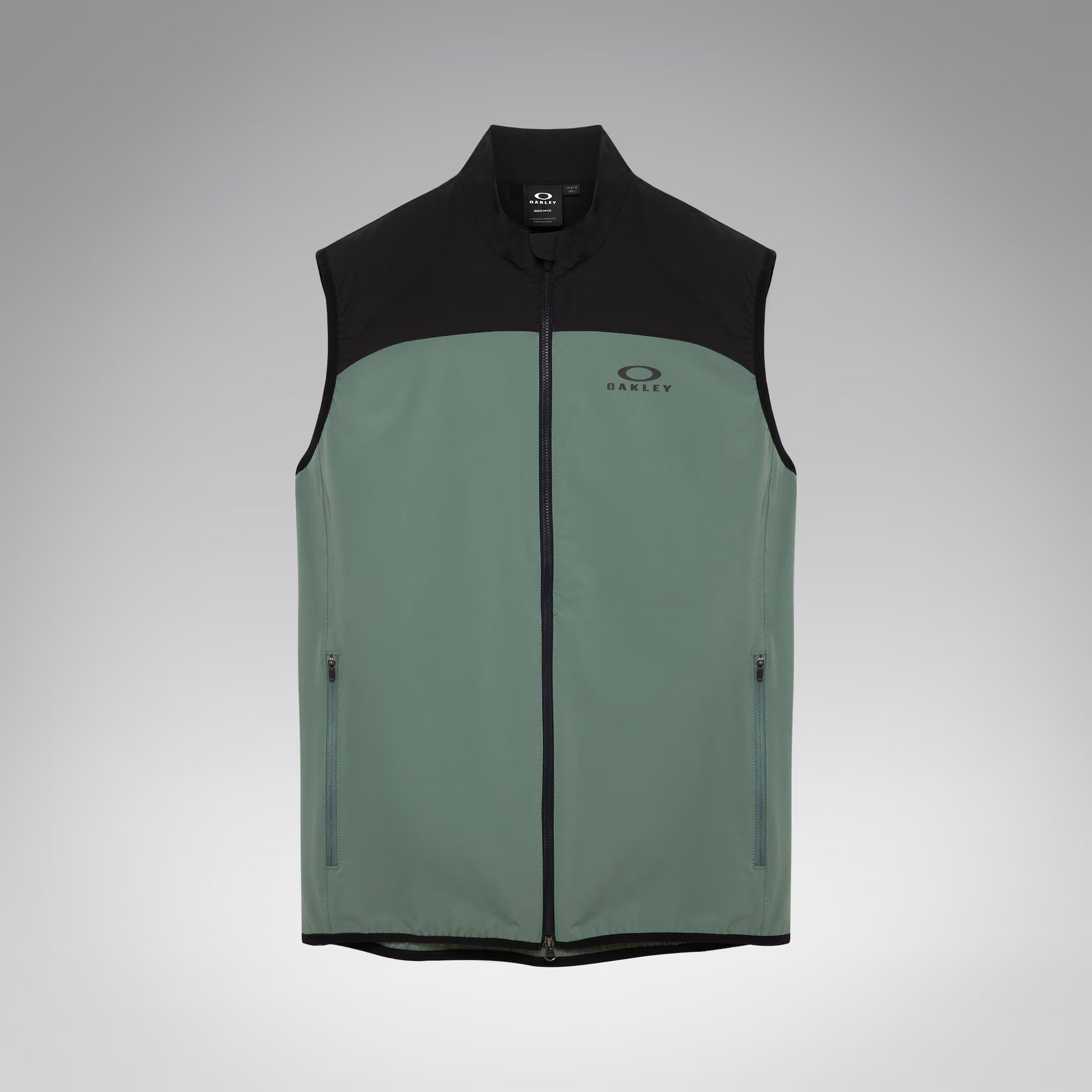 A sleeveless zip-front vest with a black upper yoke, collar, and hem, and a muted sage green lower body. The vest has a small Oakley logo on the left chest and zippered side pockets, shown on a light gray gradient background.