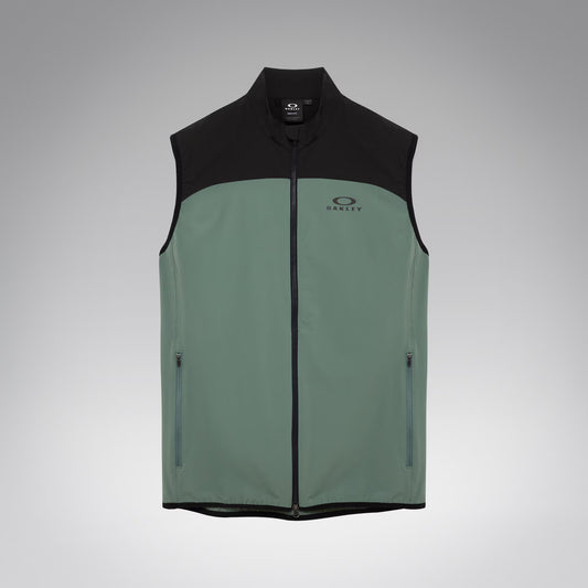 A sleeveless zip-front vest with a black upper yoke, collar, and hem, and a muted sage green lower body. The vest has a small Oakley logo on the left chest and zippered side pockets, shown on a light gray gradient background.