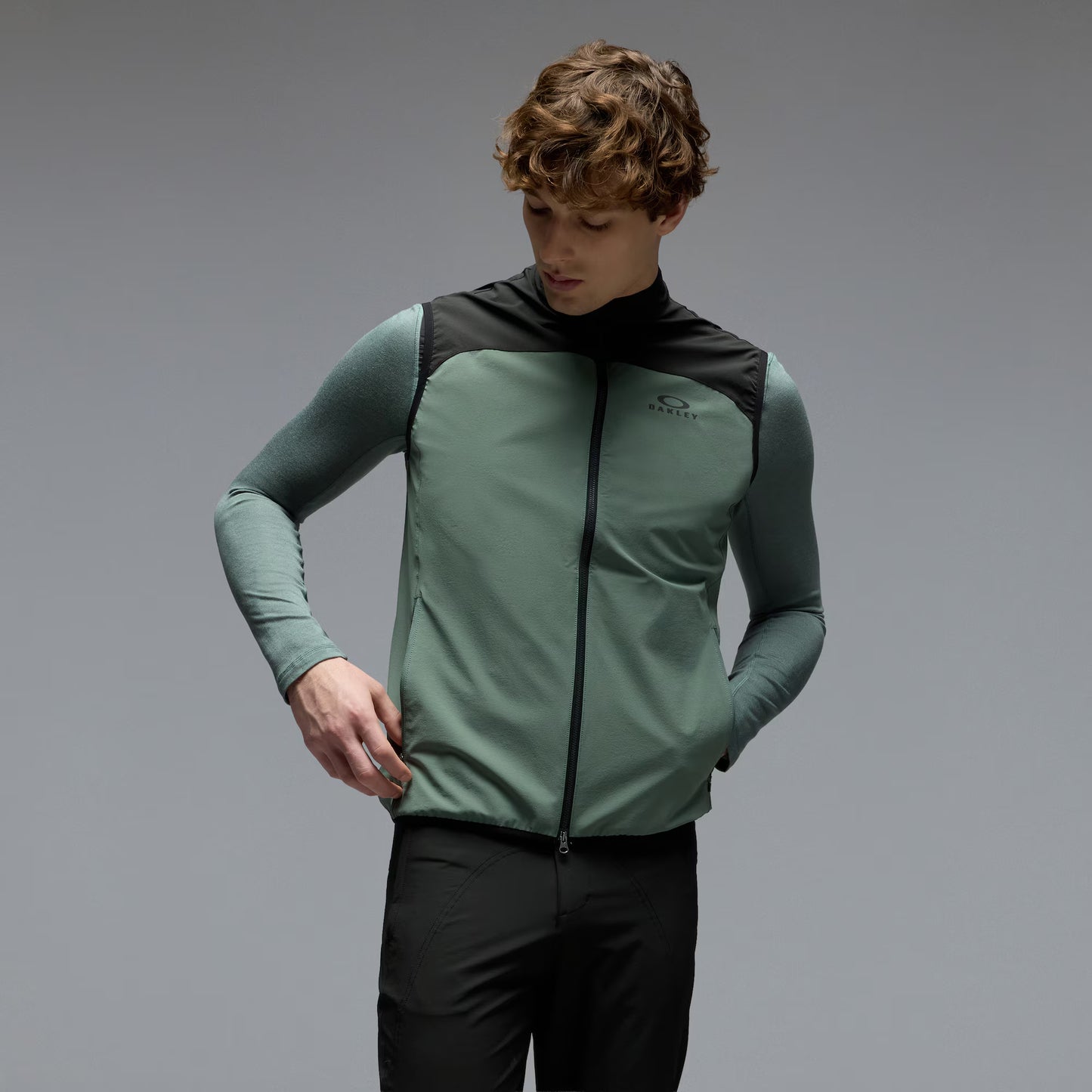 A man wearing a sage green and black athletic zip-up vest over a matching long-sleeve shirt and black pants, photographed against a neutral gray studio background.