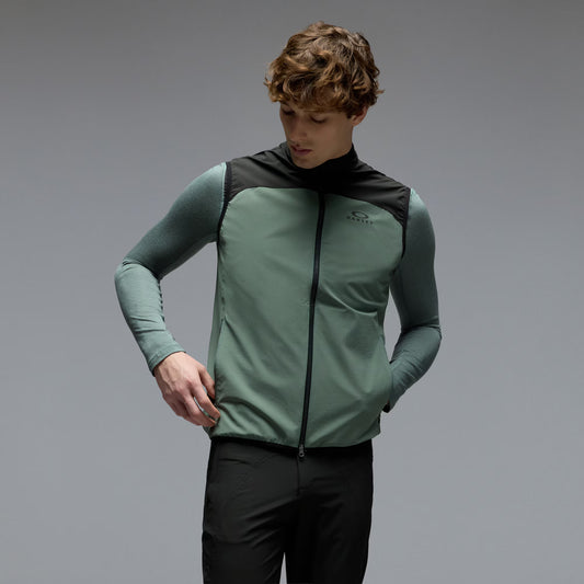 A man wearing a sage green and black athletic zip-up vest over a matching long-sleeve shirt and black pants, photographed against a neutral gray studio background.