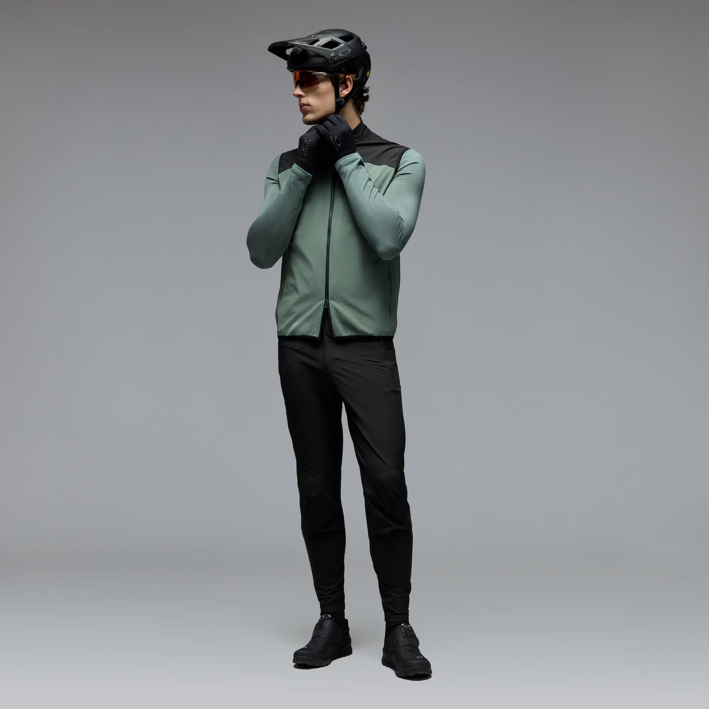 A man wearing a sage green and black cycling jersey, black cycling pants, black gloves, and a black helmet stands against a neutral gray studio background.