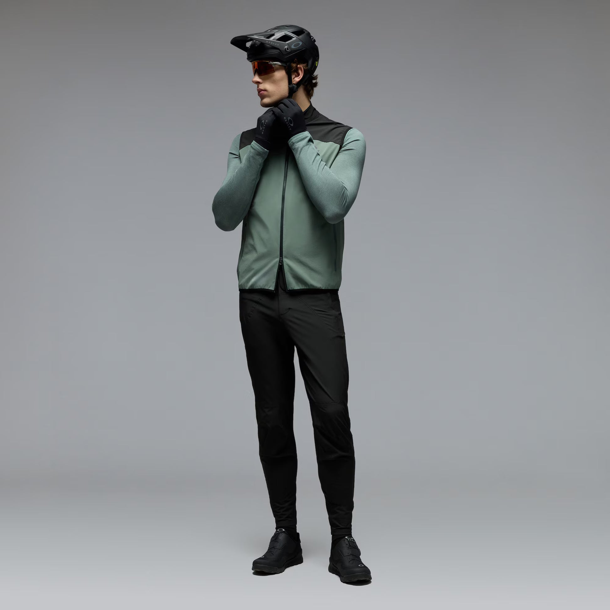 A man wearing a sage green and black cycling jersey, black cycling pants, black gloves, and a black helmet stands against a neutral gray studio background.