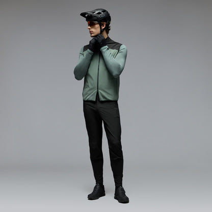 A man wearing a sage green and black cycling jersey, black cycling pants, black gloves, and a black helmet stands against a neutral gray studio background.