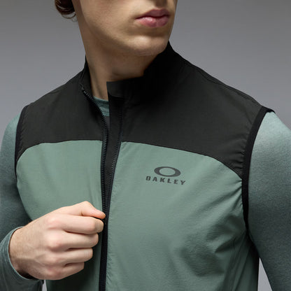 A close-up of a man wearing an Oakley sleeveless zip-front vest over a long-sleeve shirt. The vest has a black upper yoke and collar, with a muted sage green lower body and a black Oakley logo on the chest.