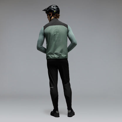 A man wearing a green and black cycling vest over a long-sleeve green shirt, black cycling pants, and a black helmet, shown from the back against a gray studio background.