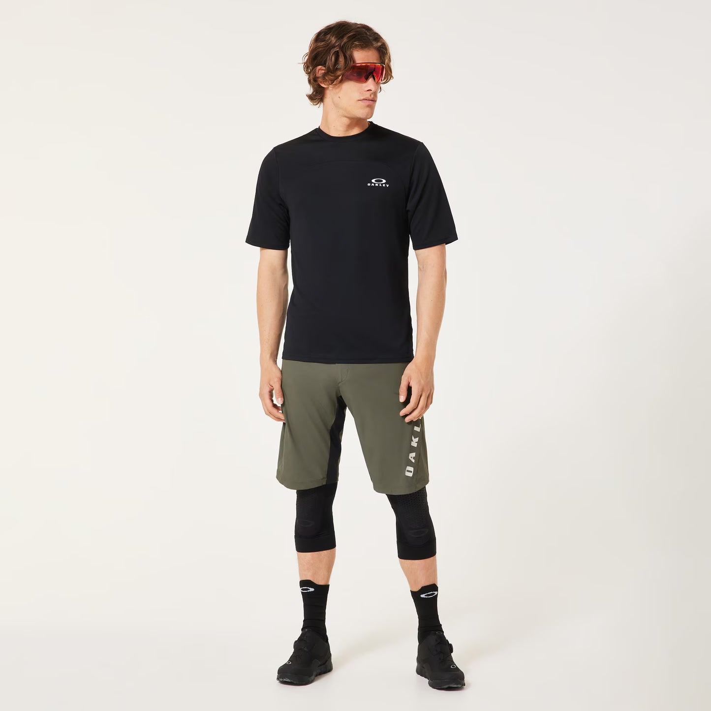 A man modeling a black short-sleeve athletic t-shirt with a small white logo on the left chest, paired with olive green shorts, black compression sleeves, black socks, and black running shoes against a plain white studio background.