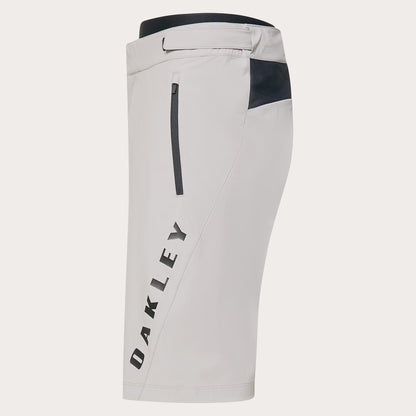 Oakley athletic shorts in light gray with a dark navy waistband, a vertical zip pocket on the left leg, a side pocket with a dark navy accent, and the Oakley logo printed vertically in dark gray on the lower left leg.