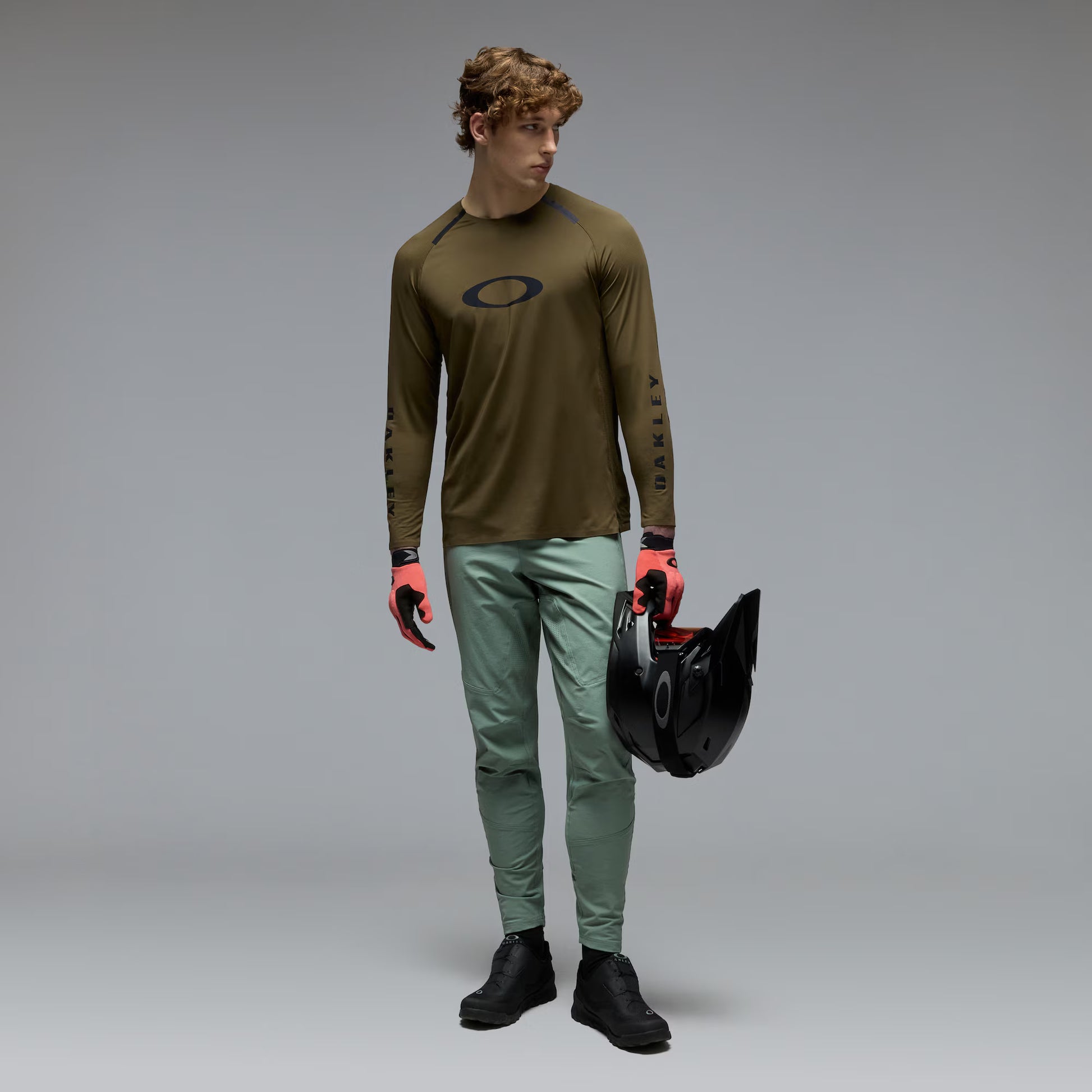 A male model wearing an olive brown Oakley long-sleeve jersey, sage green pants, and black boots, holding a black helmet against a gray studio background.
