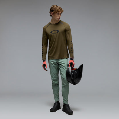 A male model wearing an olive brown Oakley long-sleeve jersey, sage green pants, and black boots, holding a black helmet against a gray studio background.