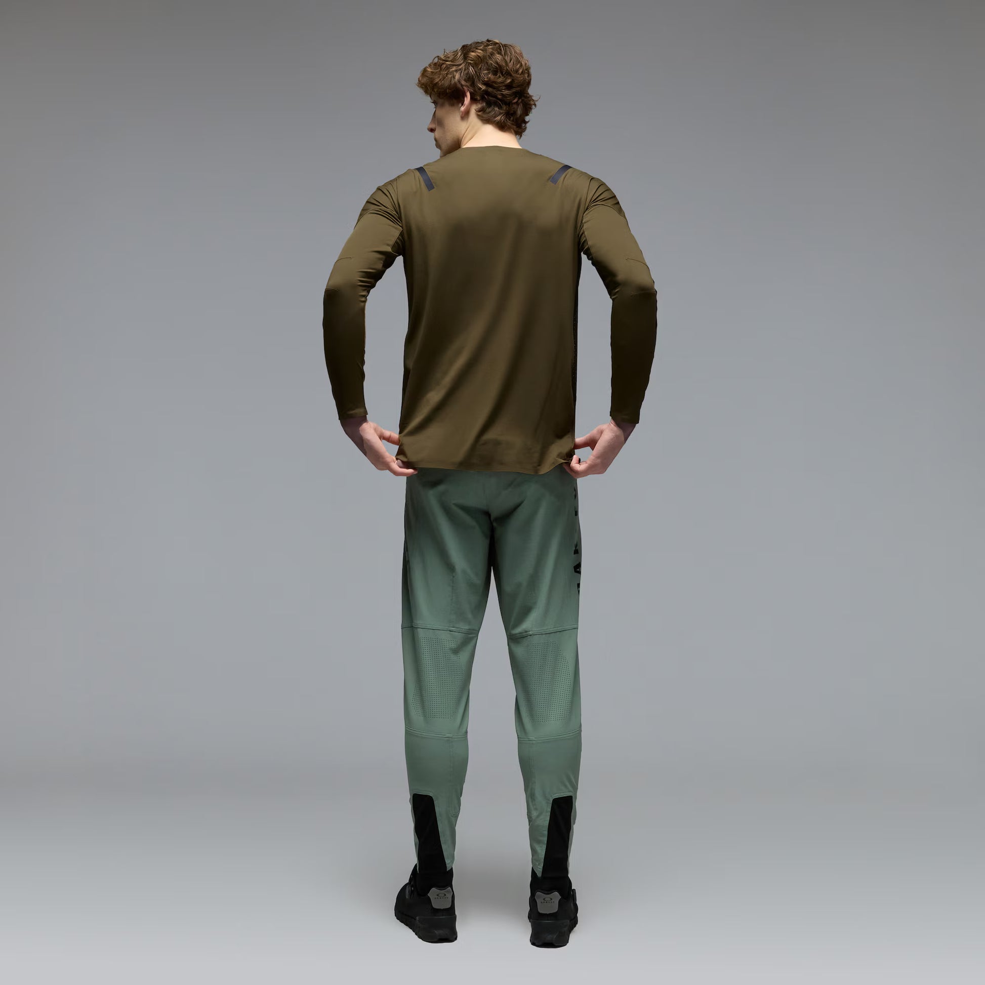 A man viewed from behind wearing an olive green long-sleeve athletic top and sage green athletic pants with black socks and black shoes, set against a neutral gray studio background.