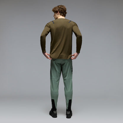 A man viewed from behind wearing an olive green long-sleeve athletic top and sage green athletic pants with black socks and black shoes, set against a neutral gray studio background.