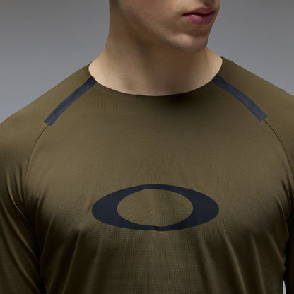 Close-up of a man wearing an olive green athletic long-sleeve shirt with a dark navy Oakley logo on the chest and dark navy shoulder panels, against a neutral gray background.