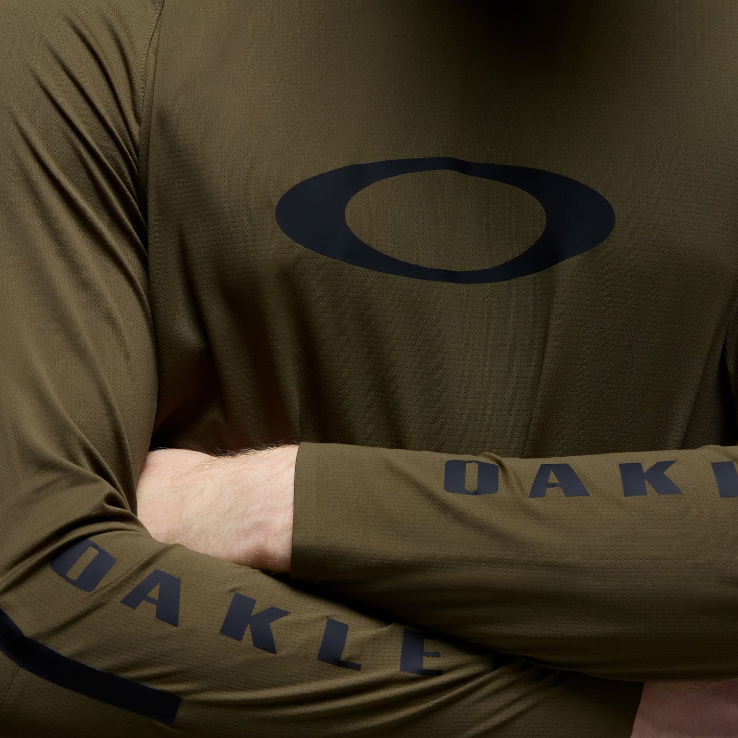Close-up of a person wearing an olive green long-sleeve athletic top with a dark navy Oakley logo on the chest and 'OAKLEY' text on the sleeve.