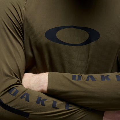 Close-up of a person wearing an olive green long-sleeve athletic top with a dark navy Oakley logo on the chest and 'OAKLEY' text on the sleeve.