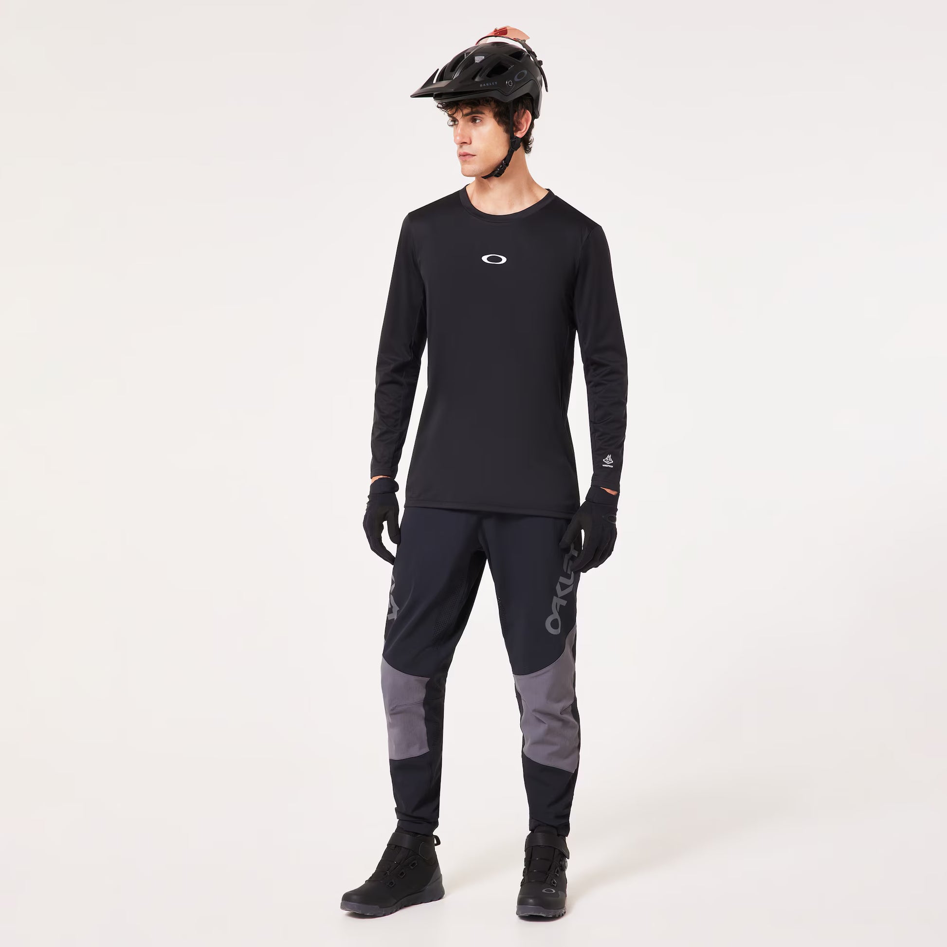 A man modeling a black long-sleeve athletic jersey with a small white Oakley logo on the chest, paired with dark gray and black cycling pants, black gloves, and a black helmet, against a plain white studio background.