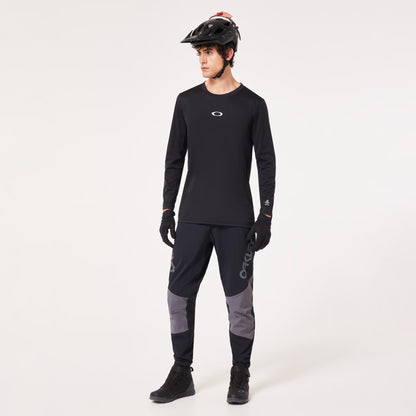 A man modeling a black long-sleeve athletic jersey with a small white Oakley logo on the chest, paired with dark gray and black cycling pants, black gloves, and a black helmet, against a plain white studio background.