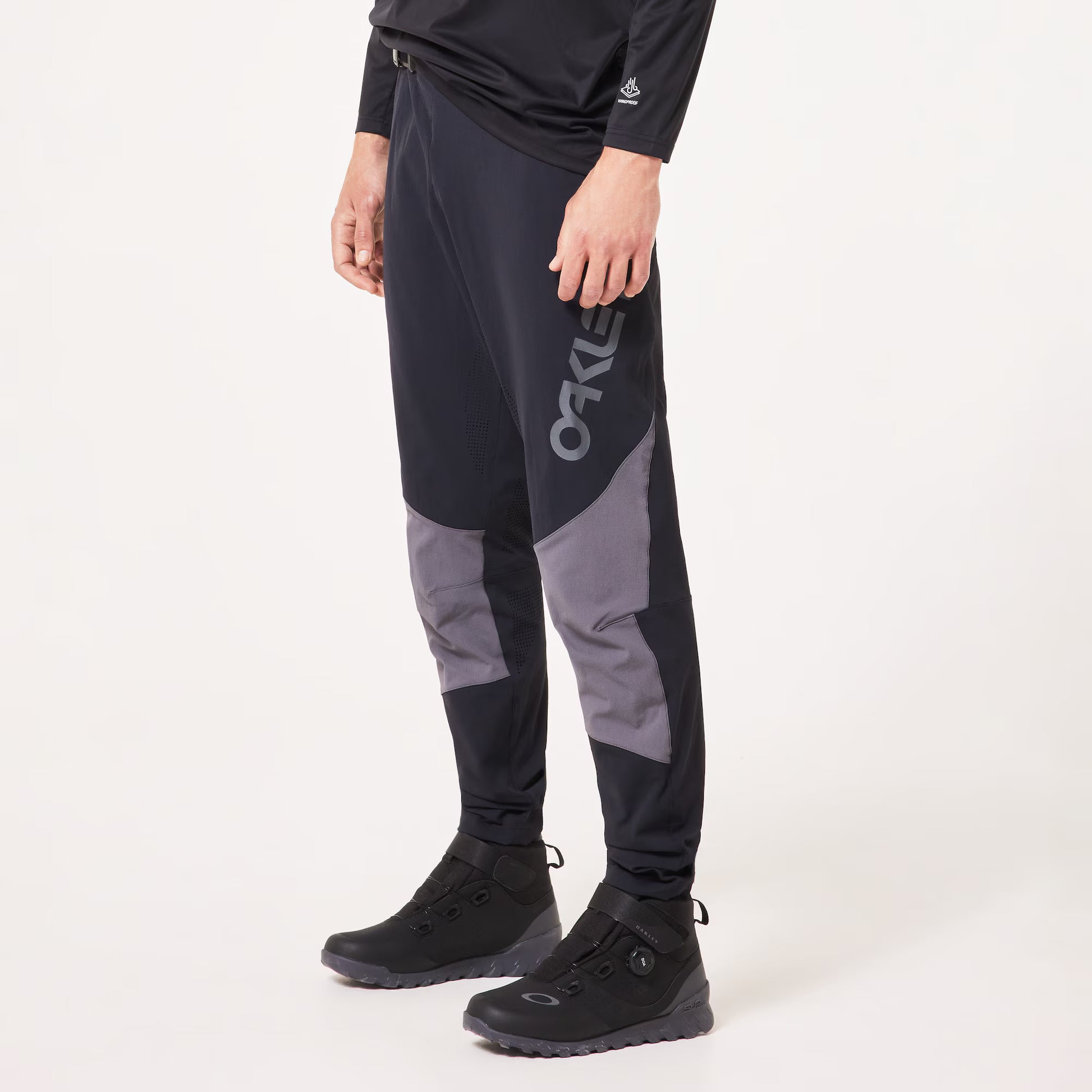A person wearing black and gray athletic jogger-style pants with a large vertical OAKLEY logo on the side, paired with black boots, photographed against a clean white studio background.