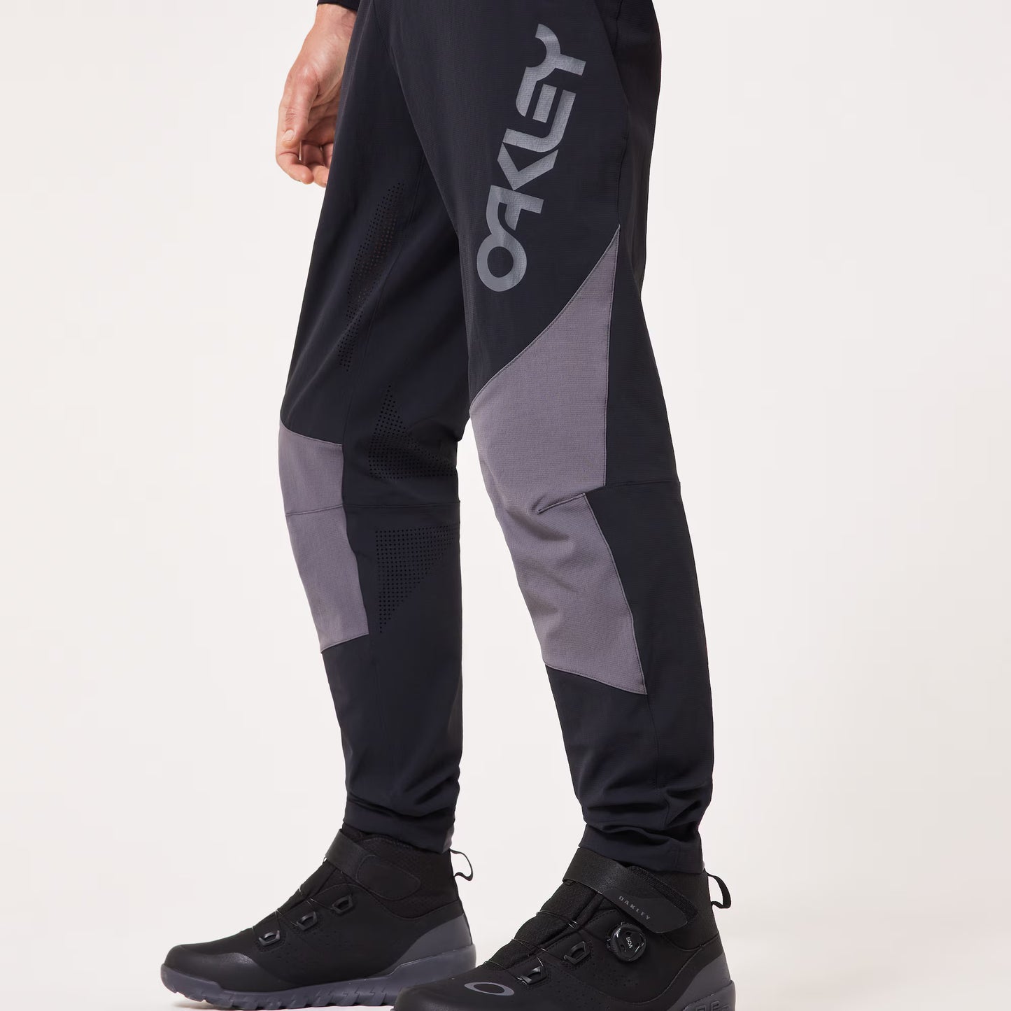 Black and gray Oakley pants worn by a model, featuring a large gray Oakley logo on the left thigh and gray knee panels, paired with black boots against a light gray background.