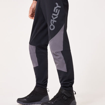 Black and gray Oakley pants worn by a model, featuring a large gray Oakley logo on the left thigh and gray knee panels, paired with black boots against a light gray background.