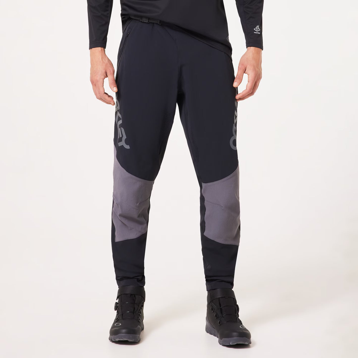 A person wearing black athletic pants with gray color-blocked knee panels and light gray logo graphics, paired with black athletic shoes, photographed against a clean white studio background.