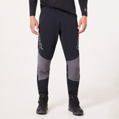 A person wearing black athletic pants with gray color-blocked knee panels and light gray logo graphics, paired with black athletic shoes, photographed against a clean white studio background.