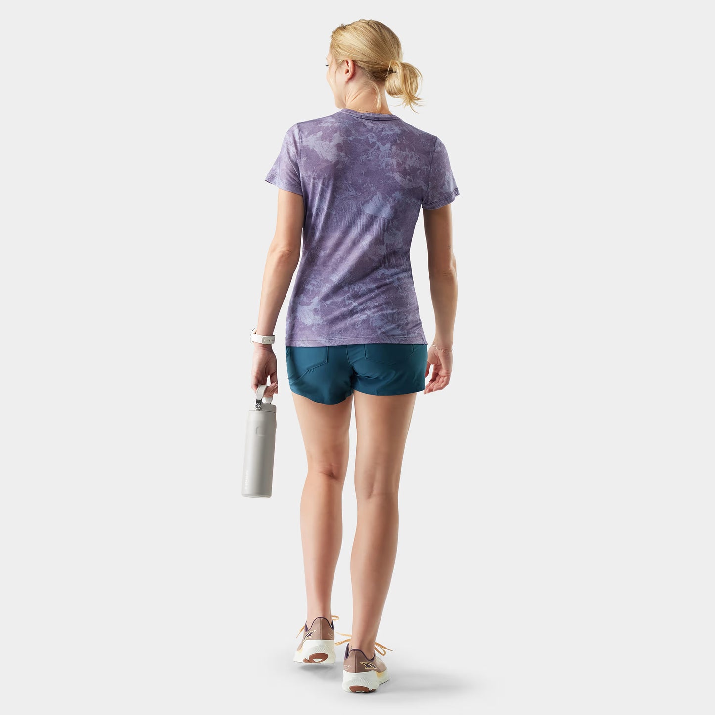 Woman wearing a purple tie-dye shirt and blue shorts, holding a white bag on a light gray background