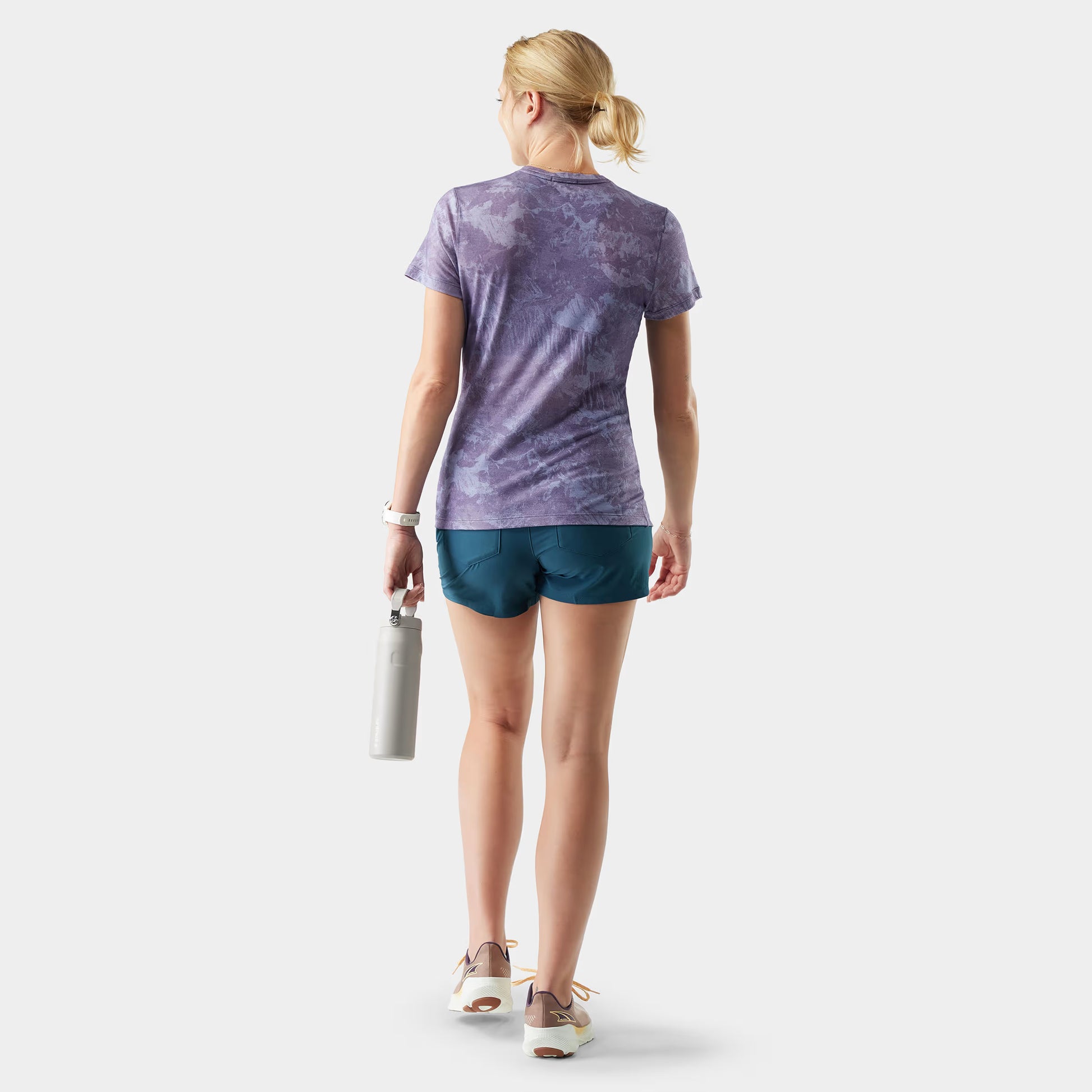 Woman wearing a purple tie-dye shirt and blue shorts, holding a white bag on a light gray background