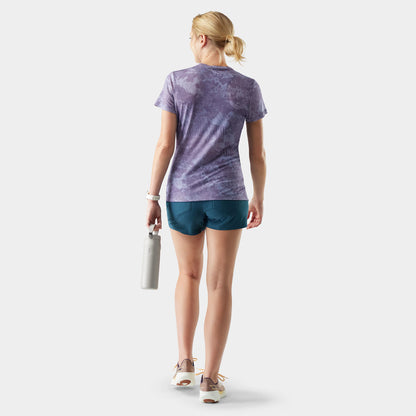 Woman wearing a purple tie-dye shirt and blue shorts, holding a white bag on a light gray background